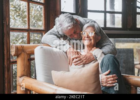 Altes kaukasisches Paar, das Freizeit zusammen zu Hause verbringt. Liebevoller Ehemann umarmt Frau von hinten auf Sessel im Haus sitzen. Ein romantischer Mann im Alter, der seinen Lebensgefährten auf einem Stuhl umarmt und sich entspannt Stockfoto