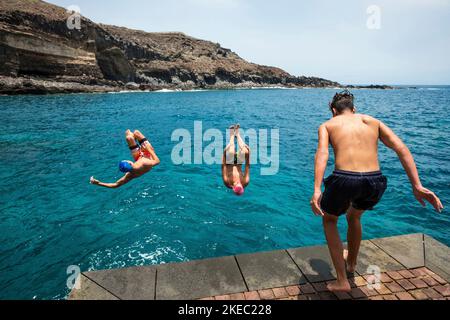 Eine Gruppe von Freunden, die zusammen am Strand abspringen, Flips machen und Spaß im Wasser haben - die Leute genießen ihren Urlaub am Strand beim Spielen und Lachen Stockfoto