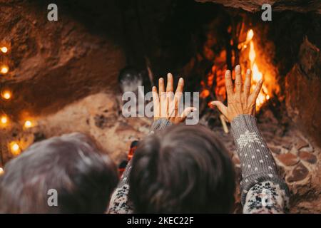 Altes kaukasisches Paar, das die Hände zu Hause warm hält. Ehemann und Ehefrau im älteren Alter entspannen sich, während sie während der Winterferien die Handfläche zum Aufwärmen am brennenden Kamin öffnen Stockfoto