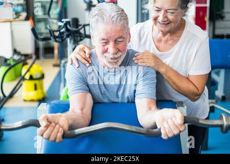 Nettes Paar von zwei seniros im Fitnessstudio, die Übungen machen - Mann mit einem barr mit Gewicht mit seiner Frau Ihm zu helfen und es zu suchen Stockfoto