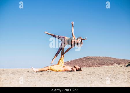 Zwei aktive und Fitness-Erwachsene, die zusammen am Strand Acro-Yoga machten - Mann lauerte auf dem Sand und hielt seine Freundin mit dem Bein Stockfoto