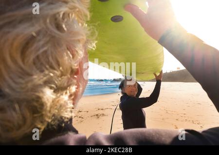 Ein paar ältere und ältere Leute oder Senioren surfen gemeinsam am Strand mit einem großen Surfbrett. Zwei Senioren, die Spaß haben und im Sommer Wassersport im Freien treiben Stockfoto