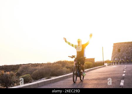 Ein reifer und Rentner genießt sein Fahrrad allein auf der Straße mit dem Sonnenuntergang im Hintergrund - Fitness und gesunde Lebensweise des alten Erwachsenen - Fahrrad fahren und Spaß mit geöffneten Armen wie Freiheitskonzept haben Stockfoto