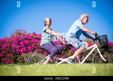 Schöne und niedliche Paar von reifen und alten Frau und Mann Reiten zusammen ein Doppel-Fahrrad in einem grünen Park mit rosa Blumen im Hintergrund. Aktiver Senior, der Spaß mit Tandem hat Stockfoto