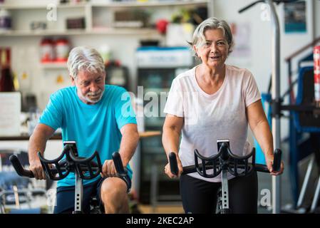 Ein paar ältere Menschen oder aktive Senioren, die im Fitnessstudio trainieren und mit dem Fahrrad in der Halle fahren, um gesund zu sein - zwei glückliche Rentner mit einem gesunden Lebensstil Stockfoto