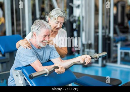 Ein paar von zwei Senioren im Fitnessstudio machen zusammen Sport Spaß haben, gesund und fit zu sein - Mann hält Ein barr ohne Gewicht und mit seiner Frau, die ihm hilft Stockfoto