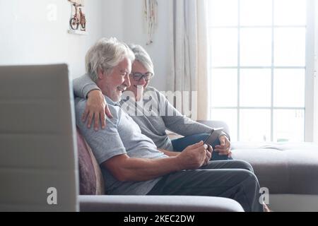Ein paar Senioren oder ältere Menschen zu Hause auf dem Sofa, die ihr Tablet oder ihr Technologiegerät lächelnd und lachend benutzen und Videos oder lustige Fotos ansehen - mobiler und technologischer Lebensstil und Konzept Stockfoto