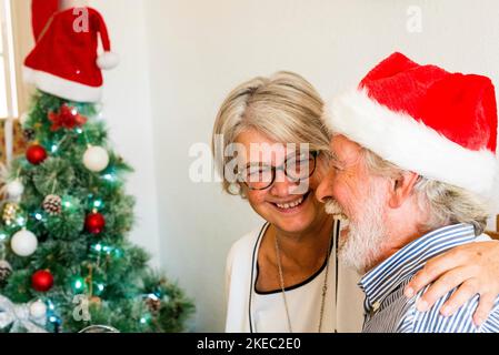 Zwei ältere Rentner, die Spaß haben und lachen Zusammen zu Hause mit einem weihnachtsbaum im Hintergrund Und mit dem reifen Mann trägt weihnachtsmütze oder Hut Stockfoto