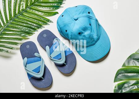 Zwei Paar blaue Flip Flops und ein Hut auf weißem Hintergrund mit Palmblättern im Vordergrund Stockfoto