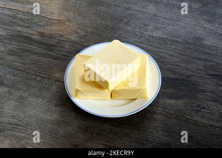 ayurvedische Ernährung, Schritte zur Zubereitung von hausgemachtem Ghee aus Butter - drei Stücke Bio-Sauerrahm-Butter auf einem weißen Porzellanteller, auf einer dunklen Holzplatte Stockfoto