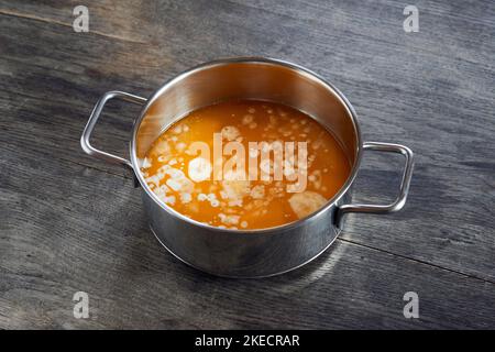 ayurvedische Diät, Schritte, um hausgemachte Ghee aus Butter vorzubereiten - geschmolzene saure Creme Butter in einem Edelstahl-Topf, auf einer dunklen Holzplatte, auf der Oberfläche beginnt Eiweiß zu setzen. Stockfoto