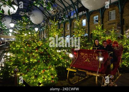 London, Großbritannien. 11.. November 2022. Zwei Frauen sitzen in einem Schlitten mit Weihnachtsbäumen hinter ihnen und lassen sich fotografieren. Der Covent Garden Markt sieht festlich aus und zieht viele Besucher an, nachdem die Lichter von Chhristmas diese Woche eingeschaltet wurden. Kredit: Elfte Stunde Fotografie/Alamy Live Nachrichten Stockfoto