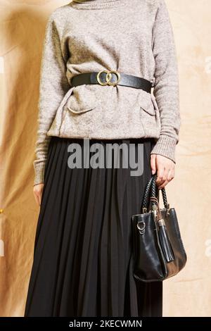 Eine Frau, die eine schwarze Handtasche hält und einen grauen Pullover mit einem goldenen Ring um den Hals trägt und vor einem steht Stockfoto