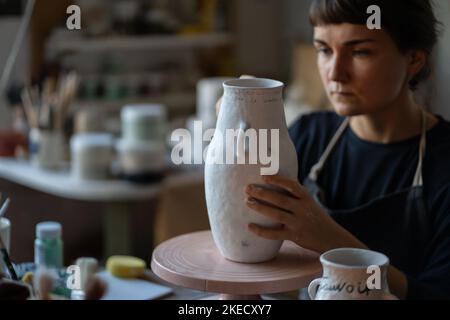 Fokussierte Brünette Frau malt Text mit Pinsel auf weiße Keramikvase am Tisch auf verschwommenem Hintergrund Stockfoto