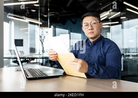 Junger asiatischer Anwalt mit Brille, der mit einem Laptop im Büro an einem Schreibtisch sitzt. Er hält einen Umschlag mit Dokumenten in der Hand. Er blickt ernsthaft in die Kamera. Stockfoto