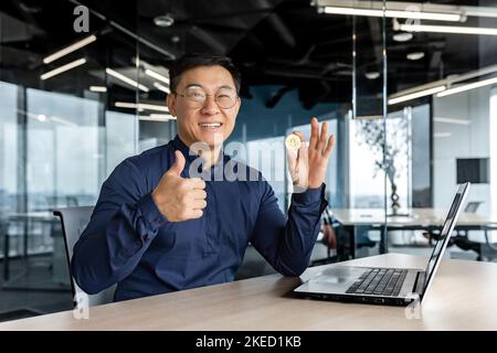 Glücklicher junger Mann asiatischer Geschäftsmann hält Bitcoin in den Händen, schaut in die Kamera, zeigt Finger super. Sitzen in einem modernen Büro mit einem Laptop, arbeiten mit Kryptowährung. Stockfoto