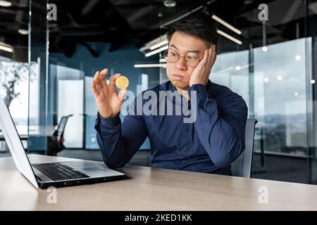 Aufgeregt und unzufrieden junger Mann Asian Geschäftsmann hält eine Bitcoin in den Händen, schaut es traurig. In einem modernen Büro mit einem Laptop sitzend, verlor man Geld bei Kryptowährungstransaktionen. Stockfoto