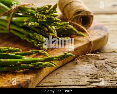 Viele frische Triebe von grünem Spargel auf einem hölzernen Hintergrund und ein Kein aus einfachem Garn. Nahaufnahme. Nützliches Produkt, gebrauchsfertig, eine Zutat für m Stockfoto