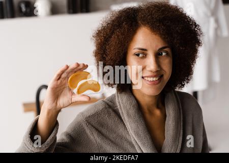 Lächelndes afrikanisches Mädchen mit goldenen Augenflecken in ihren Händen. Kosmetisches Verfahren zur Glättung von Falten, entfernen Schwellungen oder Tüten unter den Augen Stockfoto