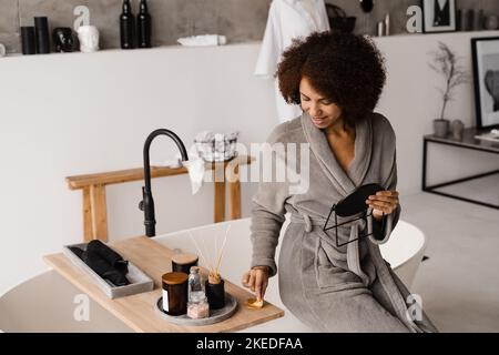 Lächelndes afrikanisches Mädchen mit goldenen Augenflecken im Bademantel im Badezimmer. Kosmetisches Verfahren zur Glättung von Falten, entfernen Schwellungen oder Tüten unter den Augen Stockfoto