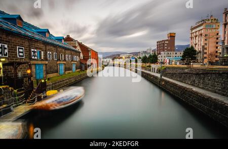 Eine lange Exposition des Himmels und des Bootes auf dem Kanal bei Otaru, Hokkaido, während der Herbst- und Wintersaison, mit Gebäuden im Hintergrund Stockfoto