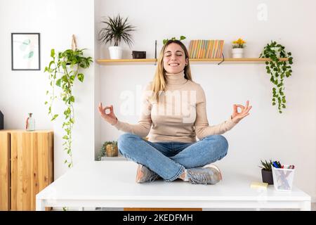 Lächelnde junge Geschäftsfrau in Lotuspose sitzt auf dem Schreibtisch im Heimbüro Stockfoto