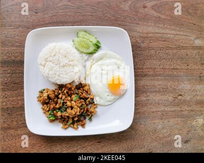 Pad kra pao gai oder rühren gebratenes Huhn mit Thai-Basilikum, Spiegelei und weißem Reis auf weißem Teller mit Holzhintergrund. Street Food in Thailand. Stockfoto