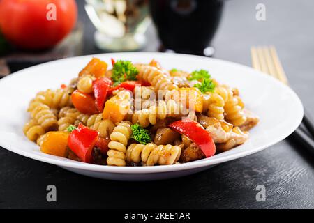 Gemüsepasta fusilli corti bucati mit Auberginen, süßem Pfeffer und Huhn in weißem Teller auf dunklem Tisch. Stockfoto