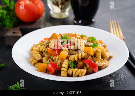 Gemüsepasta fusilli corti bucati mit Auberginen, süßem Pfeffer und Huhn in weißem Teller auf dunklem Tisch. Stockfoto