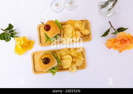 Eine obere Nahaufnahme von Burger und Chips auf den Holztellern, zwei Gläser voller Champagner, weißer Hintergrund Stockfoto