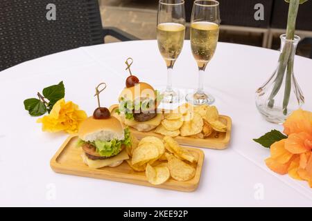 Eine obere Nahaufnahme von Burger und Chips auf den Holztellern, zwei Gläser voller Champagner, weißer Hintergrund Stockfoto