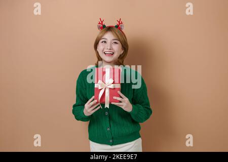 Fröhliche junge Frau in einem grünen Pullover mit Hirschgeweih hält ein Weihnachtsgeschenk und lacht Stockfoto