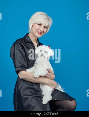 Porträt einer erwachsenen Blondine mit einem kleinen maltesischen Lapdog auf blauem Hintergrund. Stockfoto