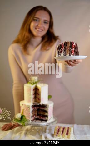 Mädchen schneidet und serviert ein Stück Kuchen. Festliche Hochzeit zweistufigen Kuchen mit frischen Blumen auf einem grauen Hintergrund dekoriert Stockfoto