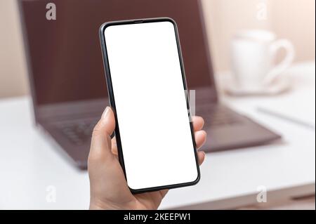 Handy auf Laptop-Hintergrund mockup. Frau Hand hält Telefon mit leerem Bildschirm im Büro. Hand hält Telefon mit weißem Bildschirm im Büro Stockfoto