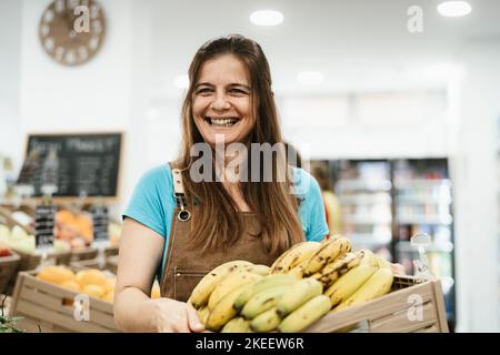 Glückliche Frau, die im Supermarkt arbeitet und eine Schachtel mit frischen Bananen hält Stockfoto