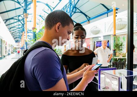 Ein Kunde, der die Kamera eines Samsung-Telefons an einem Pop-up-Verkaufsstand eines Einkaufszentrums überprüft, während ein lächelnder Mitarbeiter ihm zuschaut Stockfoto