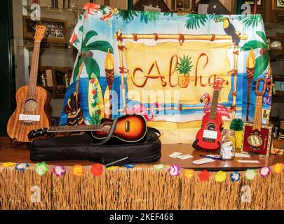 Buntes Tuch mit einer hawaiianischen Szene mit dem Wort Aloha, umgeben von einer Sammlung von Ukulelen. Stockfoto