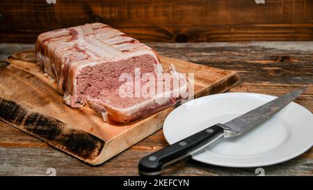 Hausgemachte Pastete in Speck aus Schweinefleisch und Leber auf rustikalem Tisch mit Messer auf dem Teller eingewickelt Stockfoto