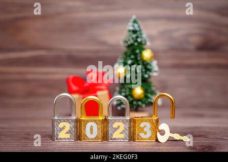 Vier glänzende Metallvorhängeschlösser, die mit 2023 Ziffern von einem Spielzeug-Weihnachtsbaum auf einem Holzhintergrund gekennzeichnet sind. Ein Vorhängeschloss wird mit einem Hauptschlüssel geöffnet. Der Koncep Stockfoto