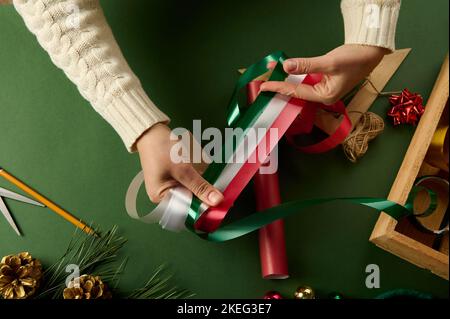 Draufsicht die Hände der Frau wählen dekorative, glänzende Bänder zum Binden von Geschenken, auf einem Hintergrund mit Verpackungsmaterialien Stockfoto