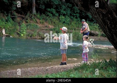 Ein kleines Mädchen sieht ihrem großen Bruder beim Fischen zu, während ihr Großvater im Hintergrund im Roaring River State Park in Missouri wirft. Stockfoto