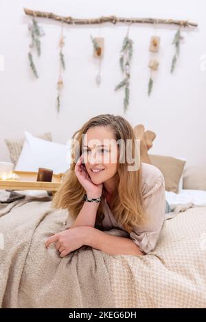 Moderne Frau mittleren Alters liegt auf dem Bett im skandinavischen Stil Weihnachten. Mädchen lächelt, trägt Pullover in neutraler Farbe, graue Hose. An einer weißen Wand, woo Stockfoto