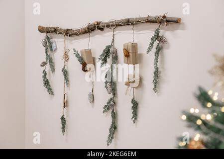 An einer weißen, hellen Wand hängt ein Holzschnäpfen, geschmückt mit Fichtenzweigen, einer Girlande, Geschenken aus Bastelpapier, Zapfen. Umweltfreundliche Dekoration für Christm Stockfoto