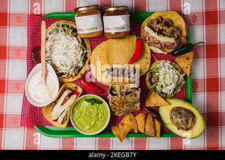 Ein Blick von oben auf ein Tablett mit verschiedenen mexikanischen Gerichten und Dips, auf einer rot karierten Tischdecke Stockfoto