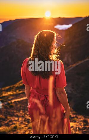 Beschreibung: Schöne braune Frau in rotem Kleid lächelt in der sonnigen Abendatmosphäre auf Pico do Ariero. Verade do Pico Ruivo, Madeira Stockfoto