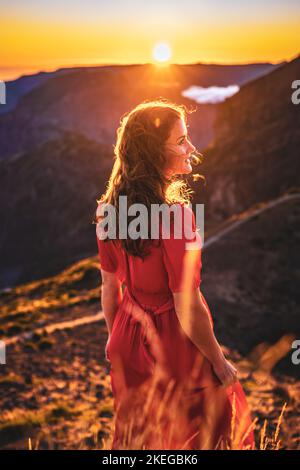 Beschreibung: Schöne braune Frau in rotem Kleid lächelt in der sonnigen Abendatmosphäre auf Pico do Ariero. Verade do Pico Ruivo, Madeira Stockfoto