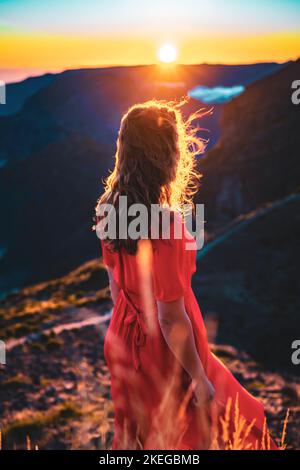 Beschreibung: Schöne braune Frau in rotem Kleid, die die sonnige Abendstimmung auf dem Pico do Ariero genießt. Verade do Pico Ruivo, Madeira Stockfoto