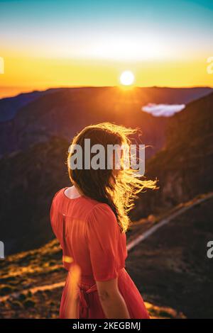 Beschreibung: Schöne braune Frau in rotem Kleid lächelt in der sonnigen Abendatmosphäre auf Pico do Ariero. Verade do Pico Ruivo, Madeira Stockfoto