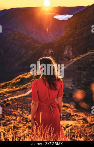 Beschreibung: Schöne braune Frau in rotem Kleid, die die sonnige Abendstimmung auf dem Pico do Ariero genießt. Verade do Pico Ruivo, Madeira Stockfoto